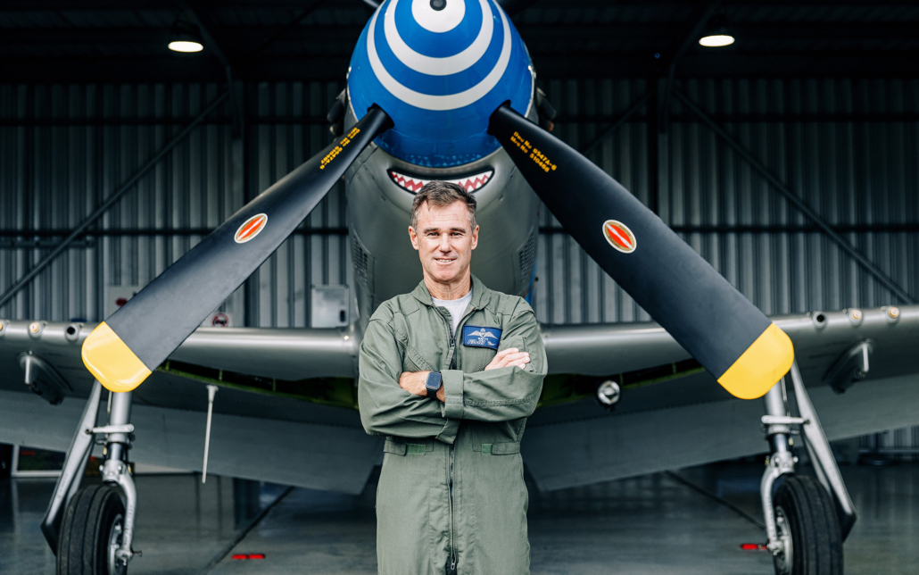 Afterburner - A man in a green flight suit stands confidently with arms crossed in front of a vintage aircraft inside a hangar, demonstrating the poise often missing when leadership development training fails. The aircraft boasts a distinctive blue nose and shark mouth artwork on its propellers.