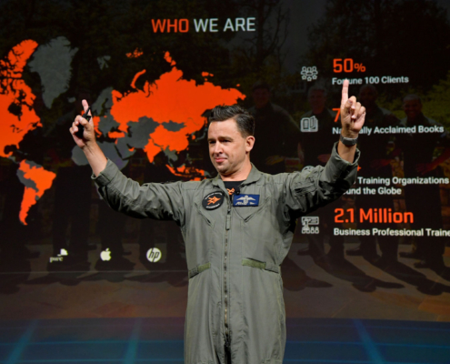 Afterburner A Person In A Flight Suit Gestures Enthusiastically On Stage, Embodying The Dynamic Energy Of An Afterburner. Behind Them, A Presentation Slide Displays Global Maps, Statistics On Business Training, And Logos Of Companies Like Pwc, Hp, And At&t.