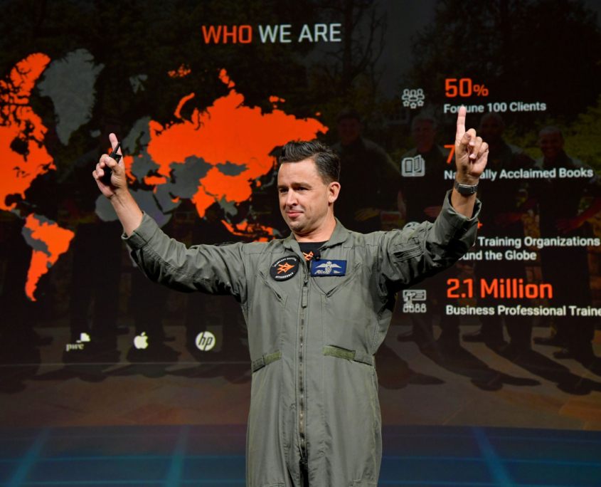 Afterburner - A person in a flight suit gestures enthusiastically on stage, embodying the dynamic energy of an afterburner. Behind them, a presentation slide displays global maps, statistics on business training, and logos of companies like PwC, HP, and AT&T. Afterburner A Person In A Flight Suit Gestures Enthusiastically On Stage, Embodying The Dynamic Energy Of An Afterburner. Behind Them, A Presentation Slide Displays Global Maps, Statistics On Business Training, And Logos Of Companies Like Pwc, Hp, And At&t.