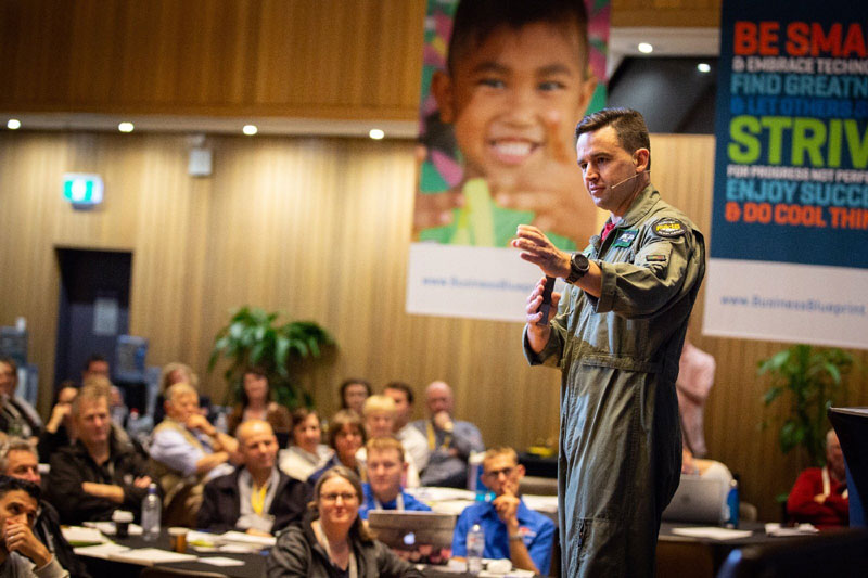 Afterburner A Speaker In A Flight Suit Presents To A Seated Audience In A Conference Hall. Behind Him, An Afterburner Banner Featuring A Smiling Child And Motivational Words Sets The Tone For An Inspiring Session.