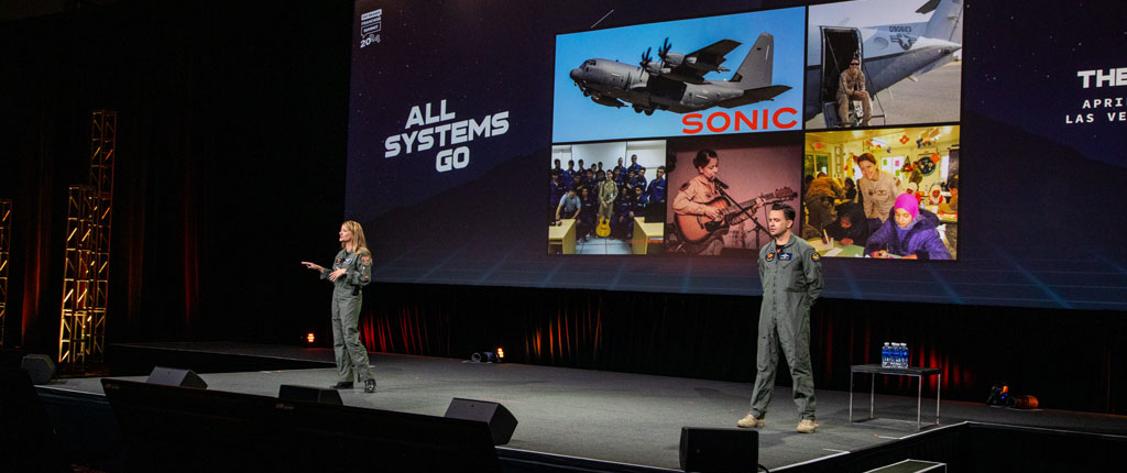 Afterburner Two Presenters In Flight Suits Take The Stage, With A Large Screen Showcasing A Military Aircraft, Musicians, And People In Costume. The Backdrop Reads All Systems Go At The Fat Brands Franchise Summit, As They Ignite The Room Like An Afterburner.