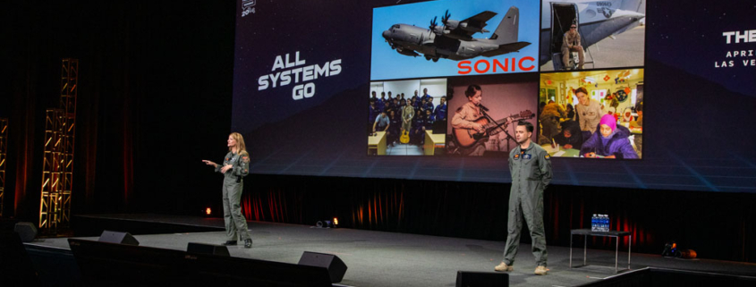 Afterburner Two Presenters In Flight Suits Take The Stage, With A Large Screen Showcasing A Military Aircraft, Musicians, And People In Costume. The Backdrop Reads All Systems Go At The Fat Brands Franchise Summit, As They Ignite The Room Like An Afterburner.